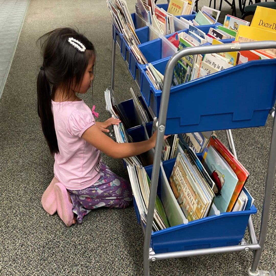 Young girl kneeling beside a rolling cart filled with children’s books, browsing titles at the Brooke Road Community Center