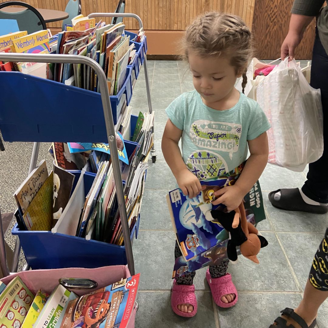 Toddler girl selecting a book from a rolling cart of children’s books at the Brooke Road Community Center