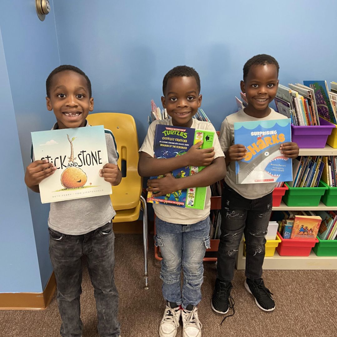 Three young boys smiling and holding books at the Brooke Road Community Center, standing in front of a bookshelf filled with children’s books