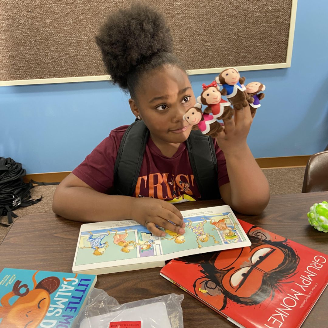 Young girl sitting at a table reading a children’s book and holding finger puppets at the Brooke Road Community Center