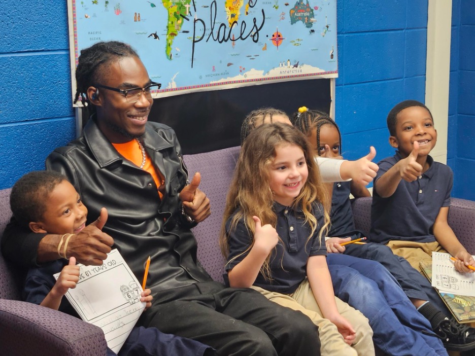 A YouthBuild volunteer sits with four smiling elementary students at Jackson Charter School, all giving a thumbs up in front of a colorful world map display.