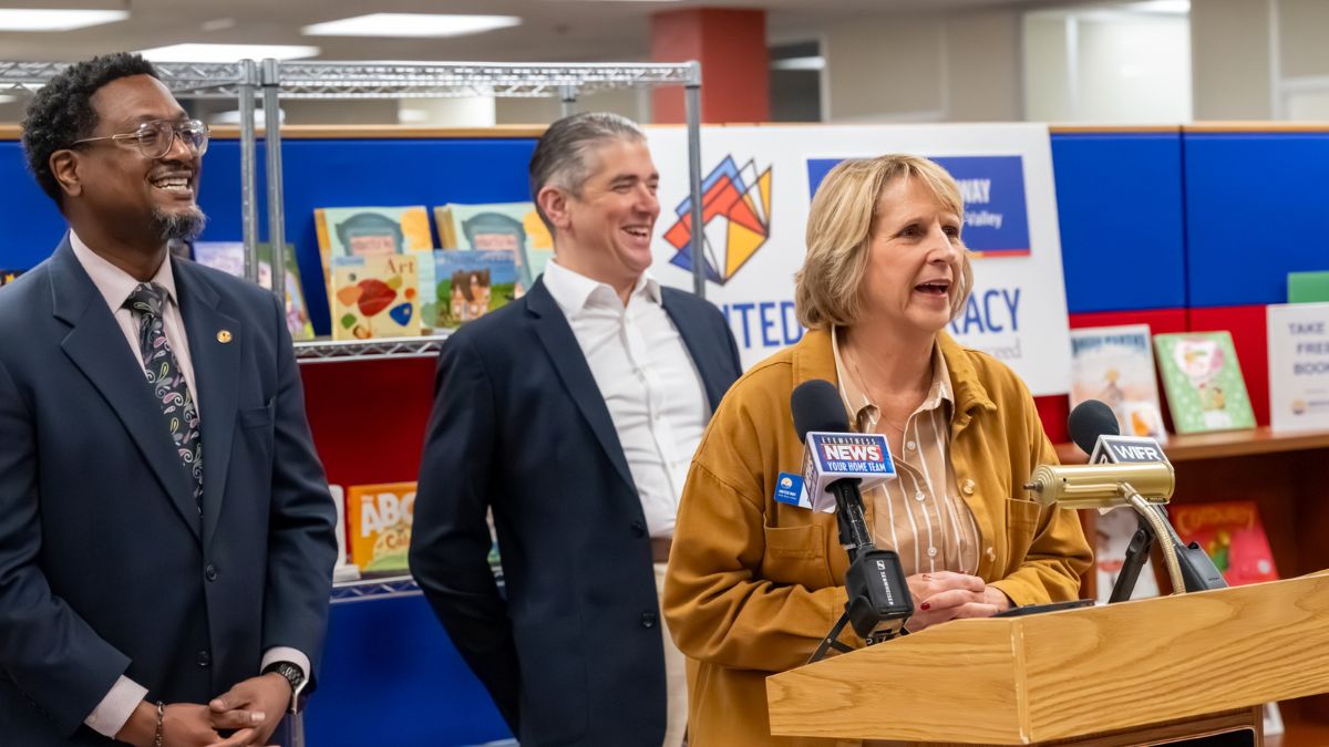 Julie Bosma, President & CEO of United Way Rock River Valley, speaks at the podium during the Books for Brighter Futures press conference, flanked by State Representatives Maurice West and Dave Vella, with children's books displayed on shelves behind them.