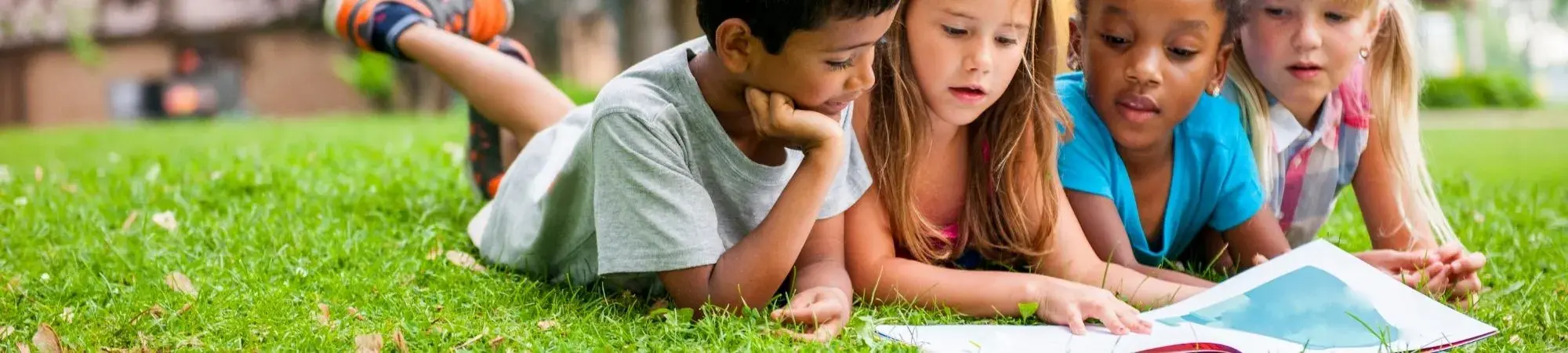 Four children lying on the grass together, reading a book outside on a sunny day.