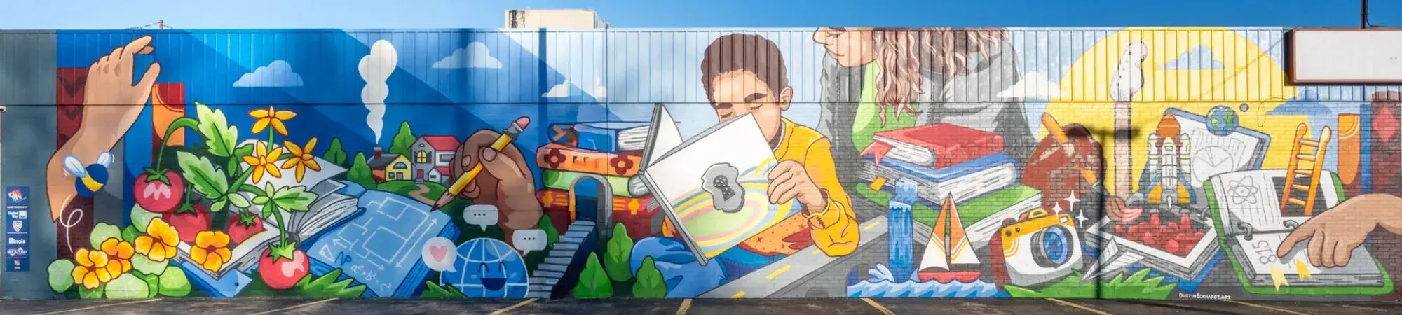 A large, colorful mural on the exterior wall of a building in Rockford, Illinois, depicting themes of literacy, community, and opportunity. The mural features a child in a yellow shirt reading a book at its center, surrounded by imagery including flowers, tomatoes, architectural blueprints, a globe, stacked books, a rocket, a camera, and figures of youth reaching toward a bright future. Painted in a bold, graphic style with vibrant blues, greens, and yellows against a bright blue sky.