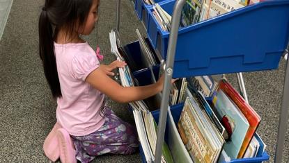 Young girl kneeling beside a rolling cart filled with children’s books, browsing titles at the Brooke Road Community Center.