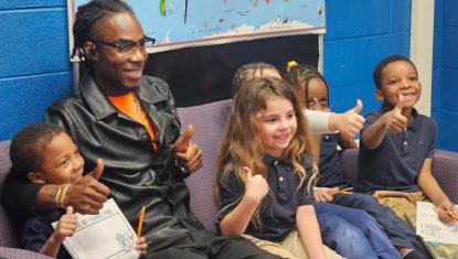2:39 PMA YouthBuild volunteer sits with four smiling elementary students at Jackson Charter School, all giving a thumbs up in front of a colorful world map display.