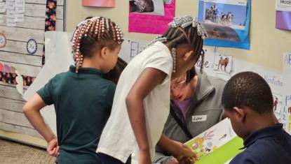A volunteer reads aloud from a picture book with three first-grade students gathered around her in a Jackson Charter School classroom.