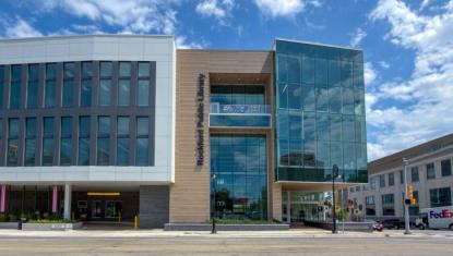 Exterior of the modern Rockford Public Library main branch, a multi-story glass and stone building with 'Rockford Public Library' signage, photographed from the street on a sunny day.